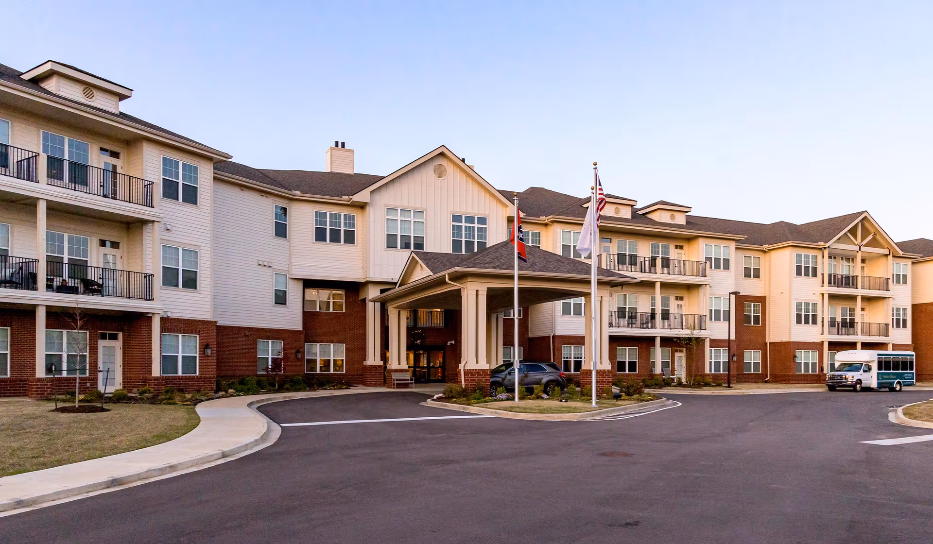 Exterior view of a multi-story senior living facility building with balconies, a covered entrance, three flagpoles with flags, a driveway, and a parked vehicle and shuttle bus.