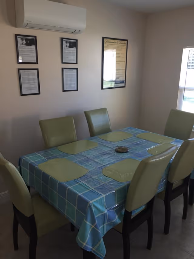 Dining room with a rectangular table covered by a blue plaid tablecloth, six green chairs and placemats, framed documents on the wall, and a wall-mounted air conditioner.
