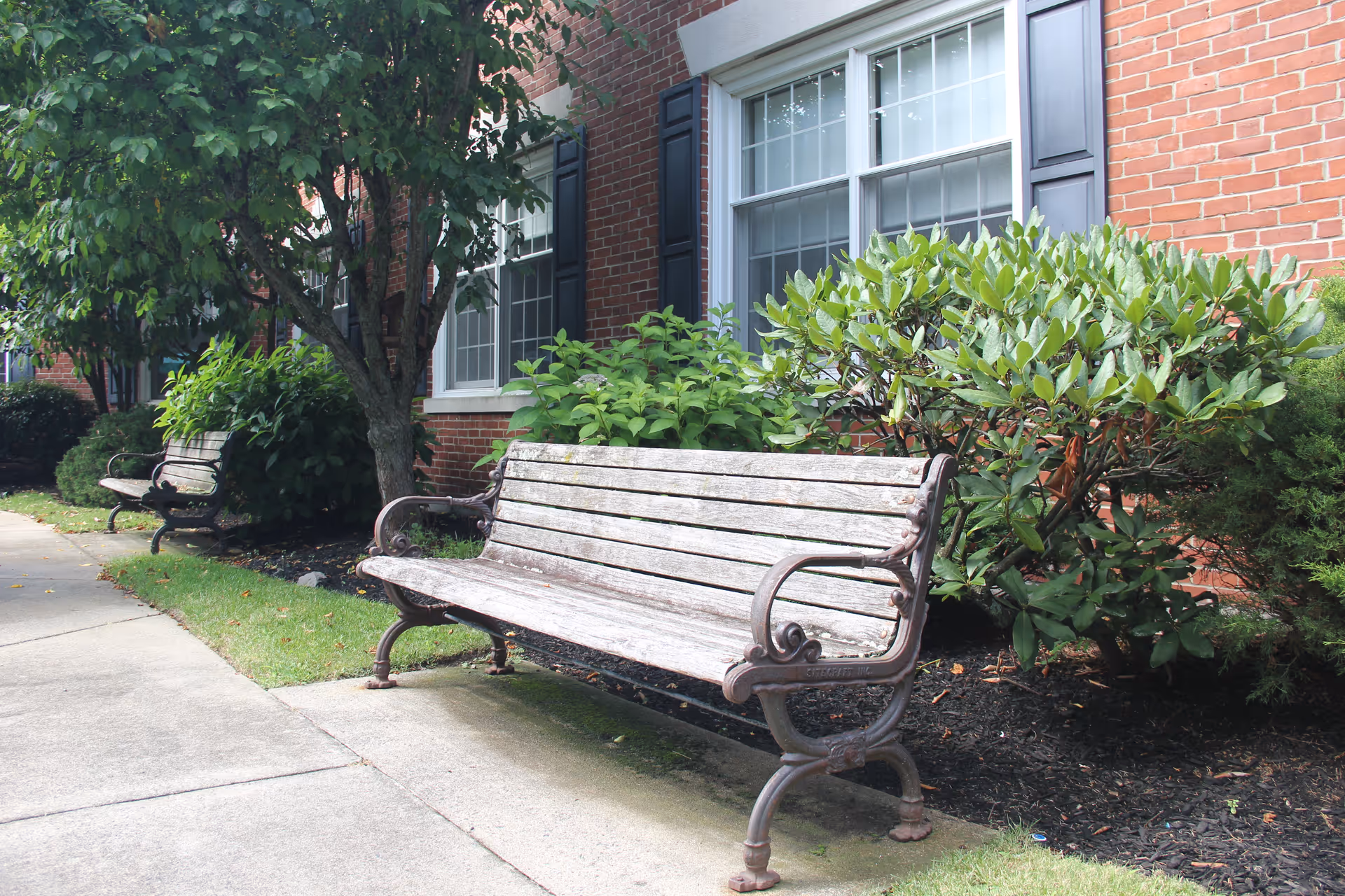 Two wooden benches with metal armrests placed on a sidewalk next to a brick building with windows and surrounded by green bushes and trees.