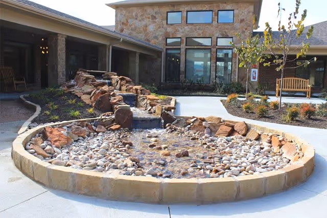 Outdoor courtyard area at Creekside Terrace Rehabilitation featuring a stone water feature with flowing water over rocks, surrounded by a concrete pathway, landscaping with small plants and trees, and a building with large windows and stone facade in the background.