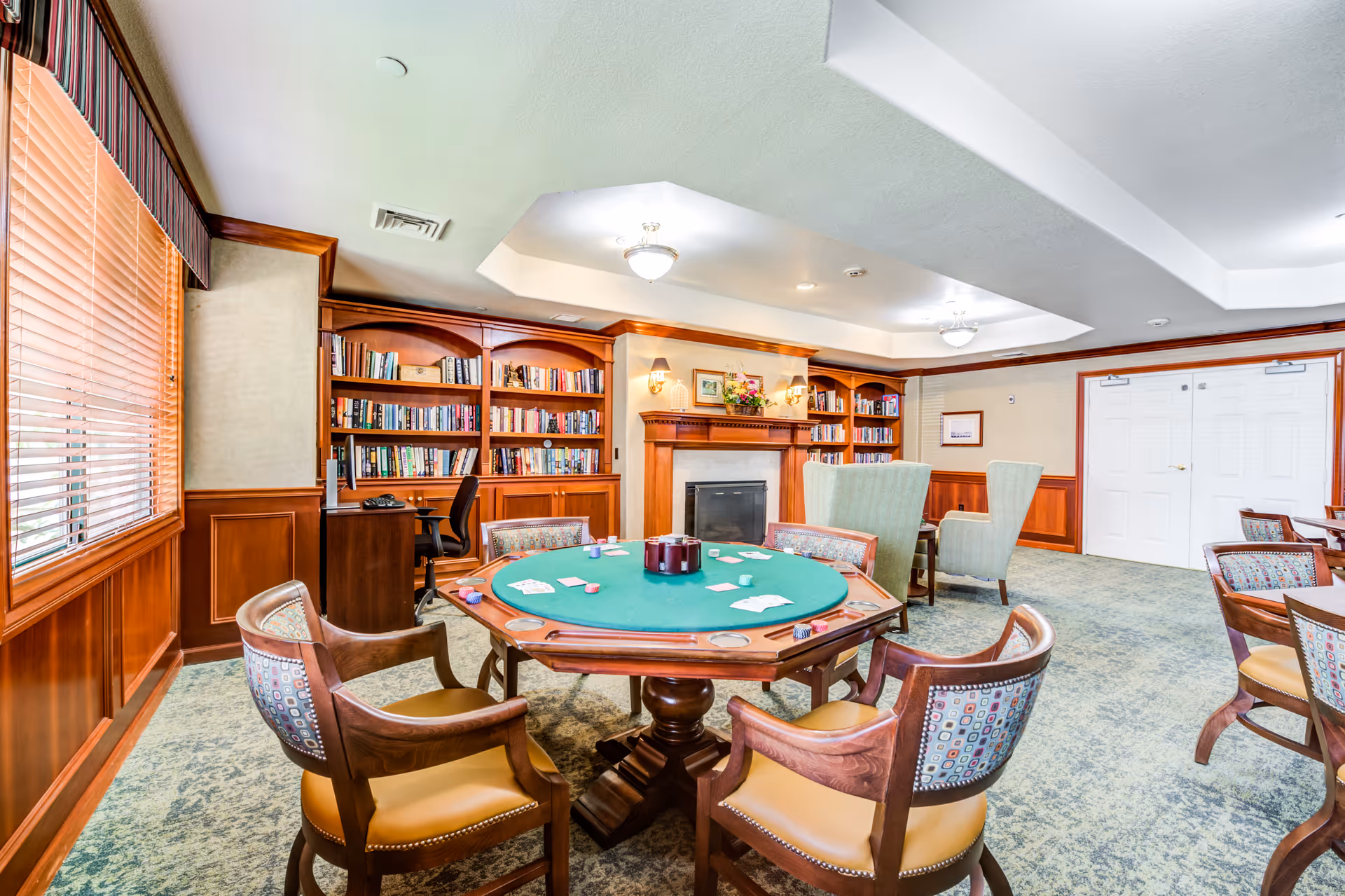A cozy senior living facility common room with a green felt poker table surrounded by wooden chairs with patterned cushions. The room features built-in wooden bookshelves filled with books, a fireplace with a decorative mantle, two high-back armchairs, and large windows with wooden blinds allowing natural light to enter.