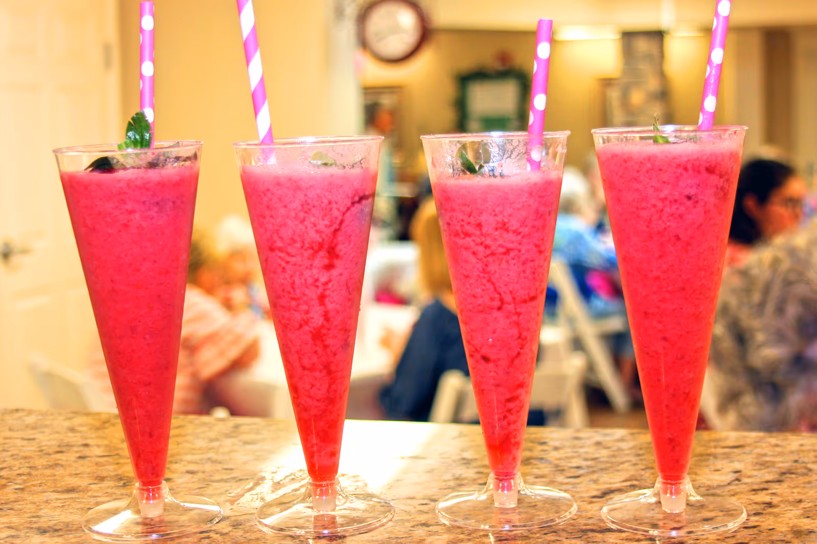 Four tall glasses filled with bright pink smoothies, each with a pink and white striped straw, placed on a countertop with a blurred background showing people sitting and socializing in a room.
