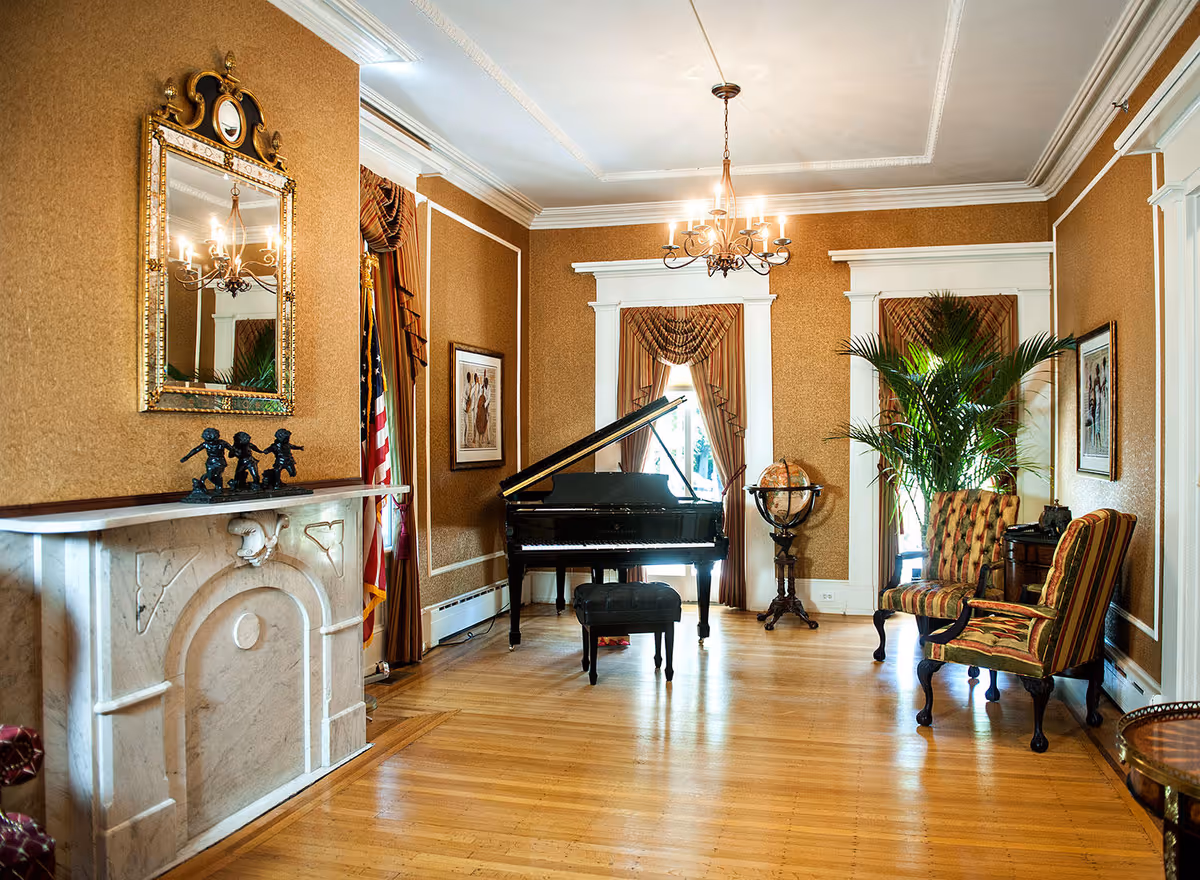 Elegant living room with a grand piano, a marble fireplace with decorative figurines, two striped armchairs, a large potted plant, framed artwork on the walls, and windows with ornate curtains. A chandelier hangs from the ceiling, and a globe stands near the window.