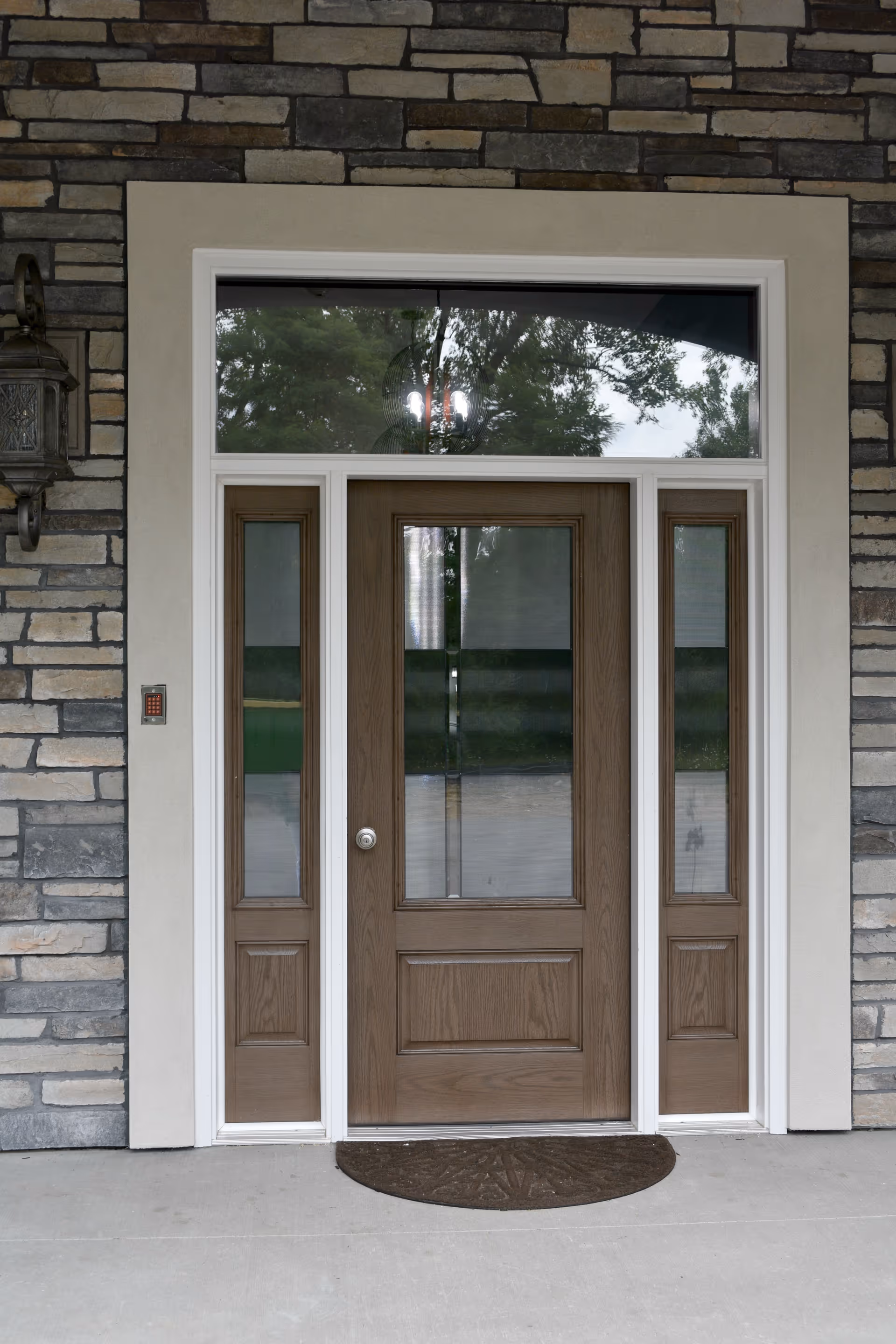 Front entrance door of a building with a wooden door featuring glass panels and sidelights, surrounded by stone and beige trim, with a brown doormat on the concrete floor and a wall-mounted lantern light on the left side.