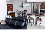 Interior view of a modern open-concept living and dining area in a retirement community. The foreground features a black leather sofa with a patterned throw pillow. Behind the sofa is a small dining table with four chairs, and to the right is a kitchen area with a breakfast bar and two high chairs. The kitchen includes stainless steel appliances and wooden cabinets. A decorative wall hanging and a chandelier light fixture are also visible.