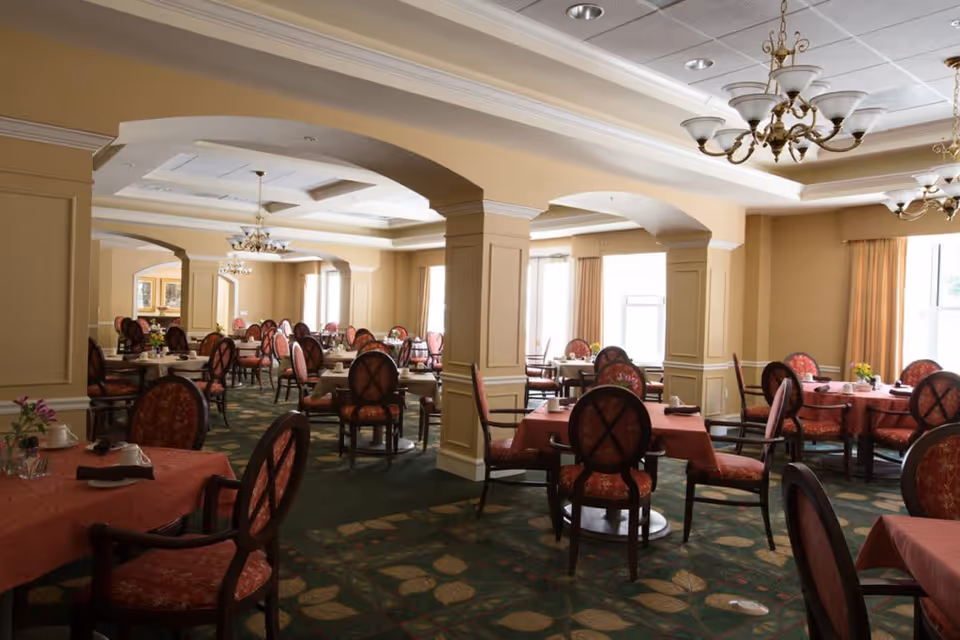 A spacious dining room with multiple round tables covered in red tablecloths, each surrounded by cushioned chairs with red patterned upholstery. The room features beige walls, large windows with curtains, decorative chandeliers hanging from a coffered ceiling, and a green carpet with leaf patterns.