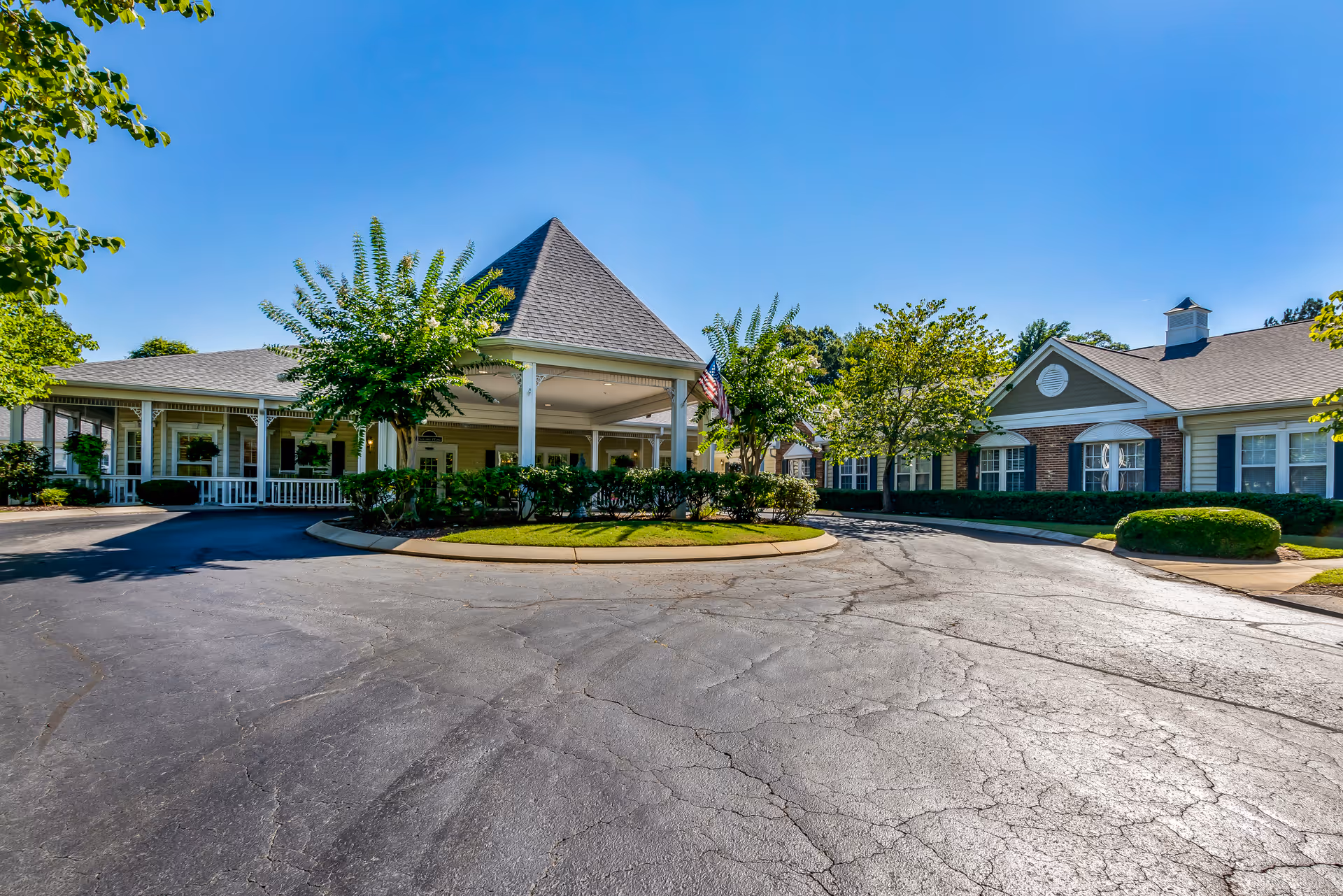 Exterior view of a senior living facility with a circular driveway, well-maintained landscaping including trees and bushes, and a covered entrance with a peaked roof under a clear blue sky.