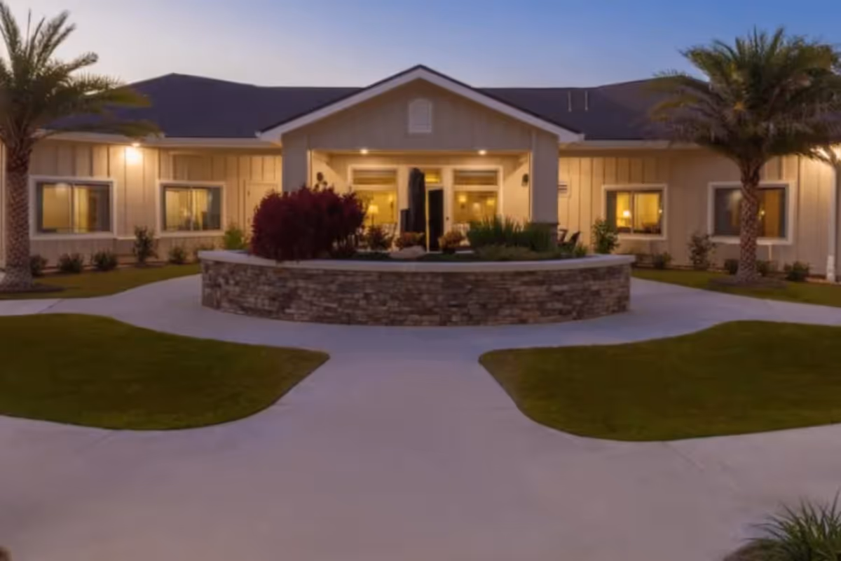 Front entrance of a one-story building at dusk with a circular stone planter, palm trees, and lit windows.