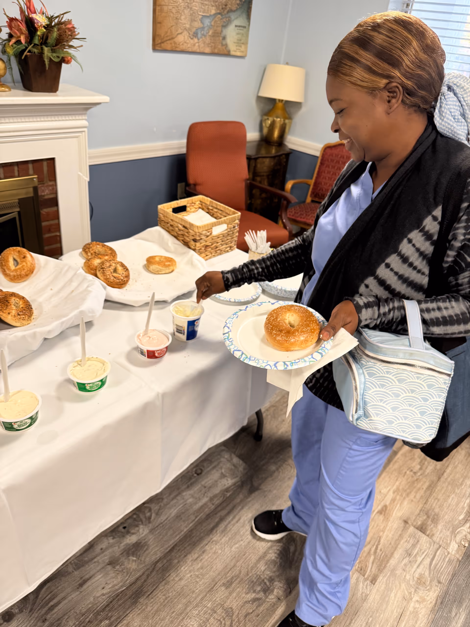 Person holding a paper plate selecting a bagel from a buffet table with bagels and spreads in a communal dining room.
