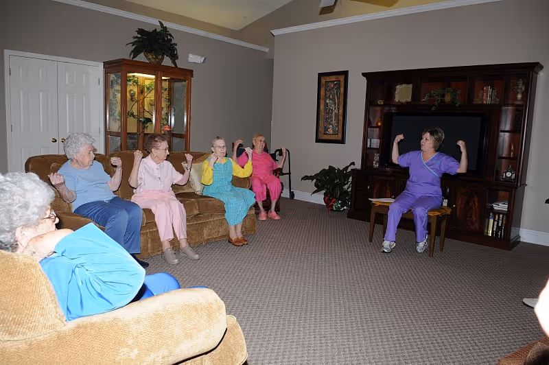 A group of elderly women sitting in a living room, following an exercise routine led by a woman in purple scrubs who is seated on a chair and flexing her arms. The room has beige walls, carpeted floor, a wooden entertainment center with a TV, and a glass display cabinet in the corner.