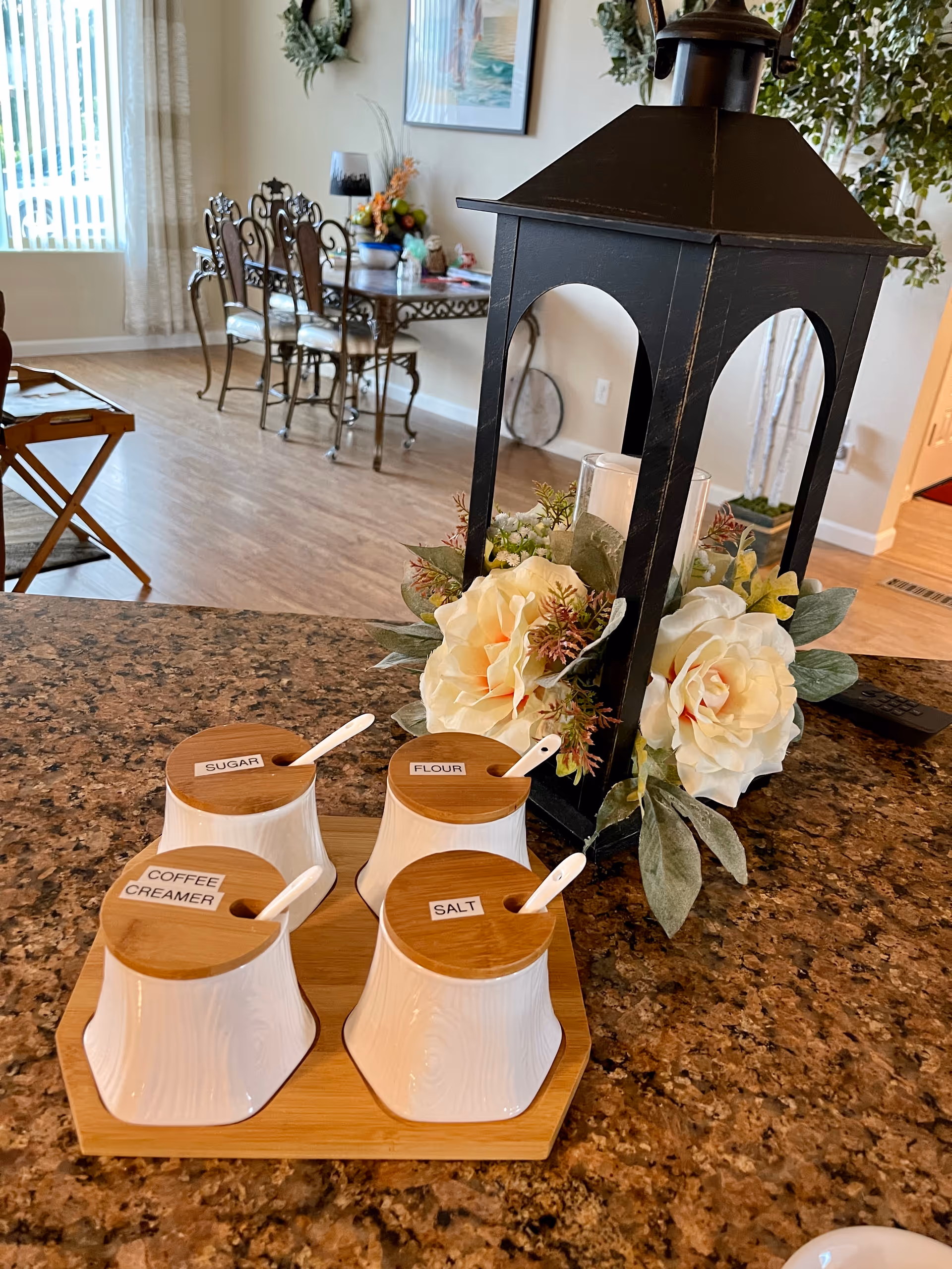 Kitchen countertop with labeled ceramic canisters for sugar, flour, salt and coffee creamer on a wooden tray beside a decorative lantern and flowers, with a dining table visible in the background.