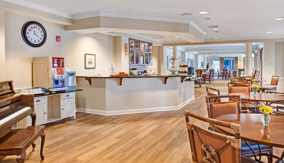 A spacious and well-lit common area with wooden floors, round wooden tables with chairs, and small vases with yellow flowers on the tables. There is a piano on the left side, a coffee machine on a white cabinet, and a counter with glass cabinets above it. The room extends into a dining area with more tables and chairs in the background.