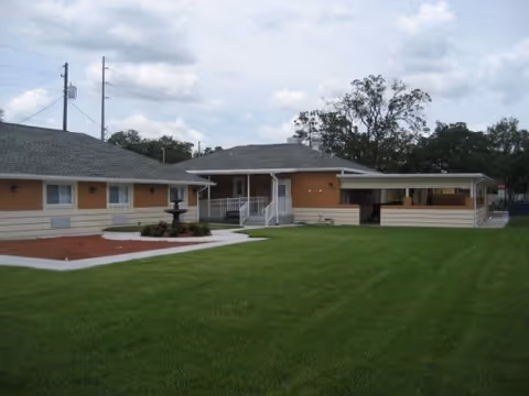 Single-story building with beige and light orange exterior walls, a gray shingled roof, and a well-maintained green lawn in front. There is a small black fountain surrounded by mulch and plants near the center walkway leading to the entrance. Trees and power lines are visible in the background under a cloudy sky.