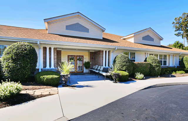 Front exterior view of a single-story senior living facility building with a covered entrance, brick and siding facade, neatly trimmed bushes, and a paved driveway under a clear blue sky.
