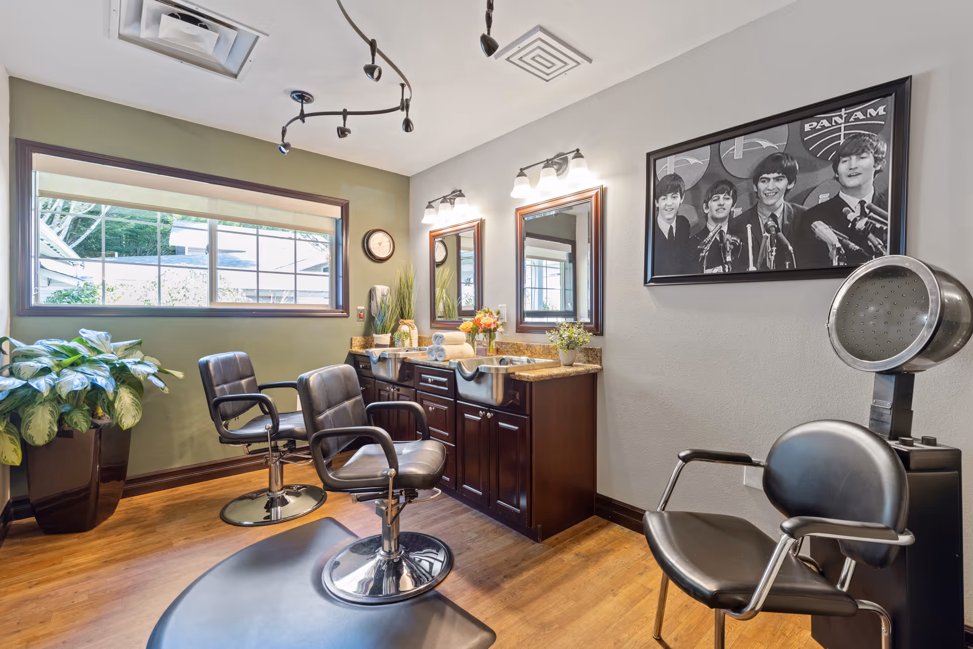 Interior of a salon room with two black salon chairs in front of a counter with two mirrors and sinks. A third black chair with a hair dryer hood is on the right. There is a large window on the left wall, a potted plant below it, and a black and white framed photo of The Beatles on the right wall.