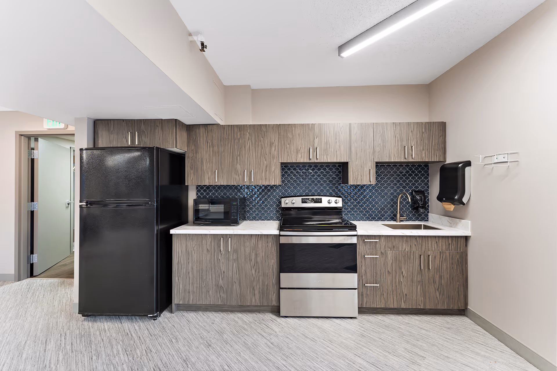 Small modern kitchen with wood-tone cabinets, a stainless steel stove, black refrigerator, microwave, sink, and blue tiled backsplash.