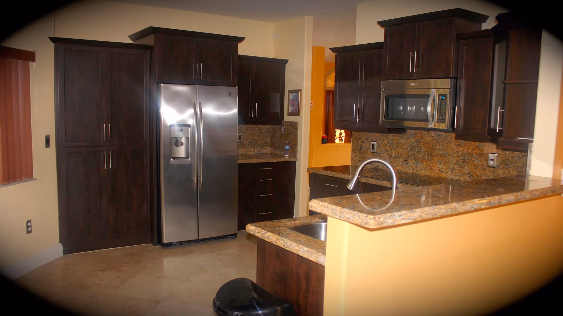 Modern kitchen with dark wood cabinets, granite countertops, a stainless steel refrigerator, built-in microwave, and a sink with a curved faucet. The floor is tiled and there is a window with vertical blinds on the left side.