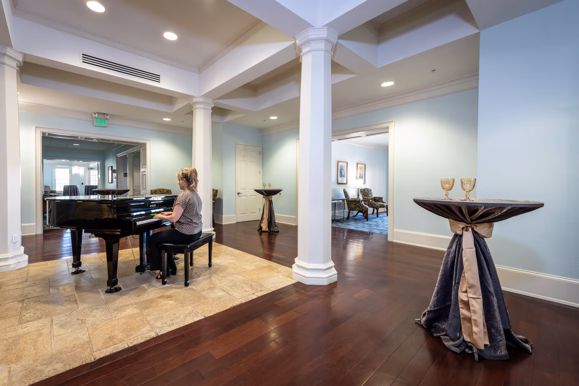 A woman playing a black grand piano in a spacious room with light blue walls, white columns, and dark wood flooring. There are two small round tables draped with dark cloth and beige ribbons, each holding two goblets. In the background, there is a seating area with chairs and framed artwork on the walls.