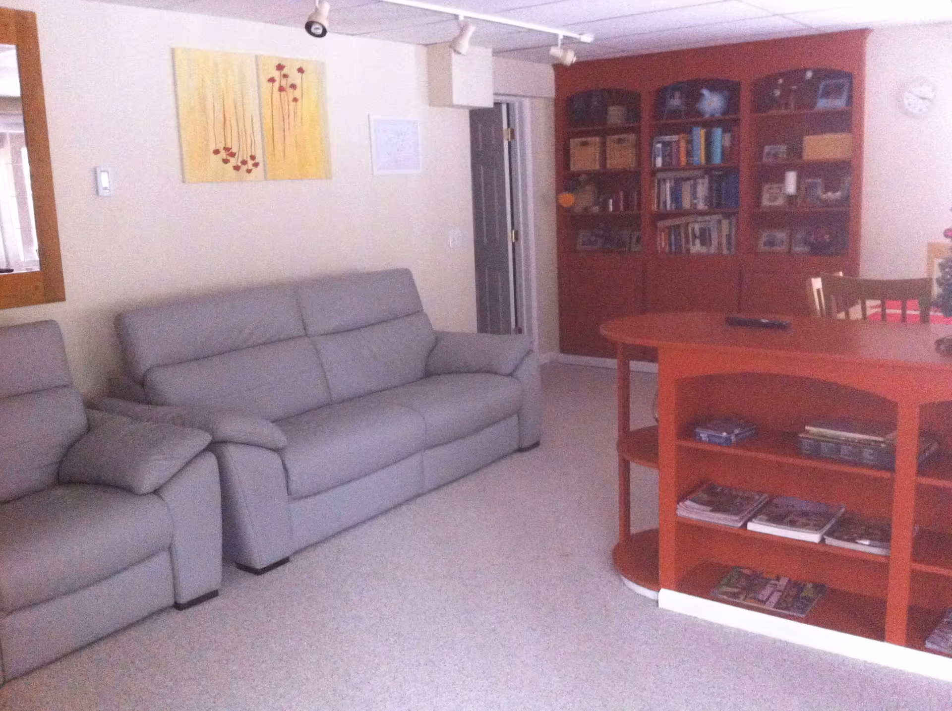 A cozy living room with a gray sofa and matching armchair, a red wooden bookshelf filled with books and decorative items, and a red wooden table with shelves holding magazines and books. The walls are light-colored with a yellow floral painting and a clock. There is a door in the background and a mirror on the left wall.