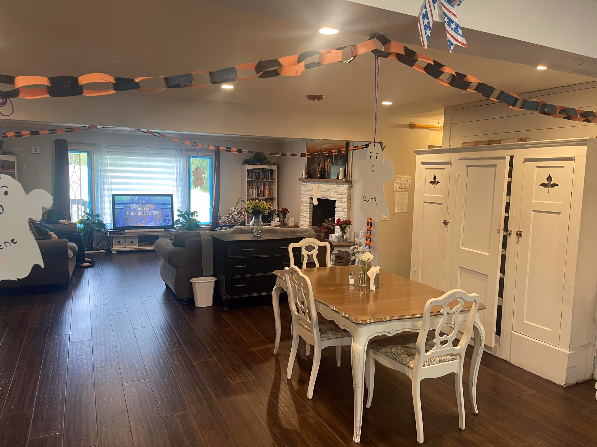 A cozy living and dining area decorated with Halloween-themed paper chains and ghost cutouts hanging from the ceiling. The room features a wooden dining table with four chairs, a dark wood sideboard, a fireplace with flowers on the mantel, a TV on a stand, and comfortable sofas near large windows with sheer curtains letting in natural light.