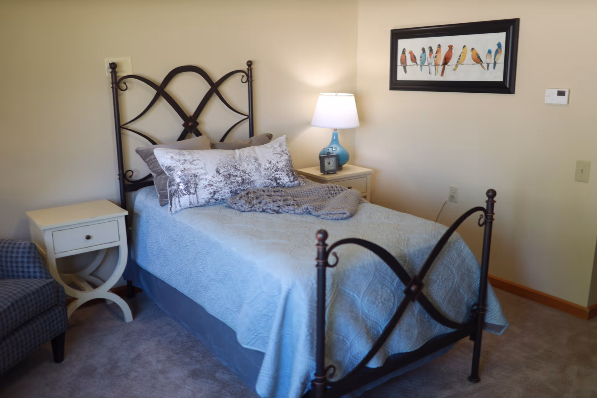 A neatly made single bed with a decorative metal headboard and footboard in a cozy bedroom. The bed has a light blue quilt, patterned pillows, and a folded gray blanket. Next to the bed is a white nightstand with a blue lamp and a small clock. A framed picture of colorful birds on a wire hangs on the beige wall above the nightstand. There is a patterned armchair partially visible on the left side of the image.