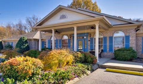 Exterior view of a single-story brick building with a covered entrance supported by white columns. The building has blue shutters on the windows and is surrounded by well-maintained bushes and flowering plants under a clear sky.