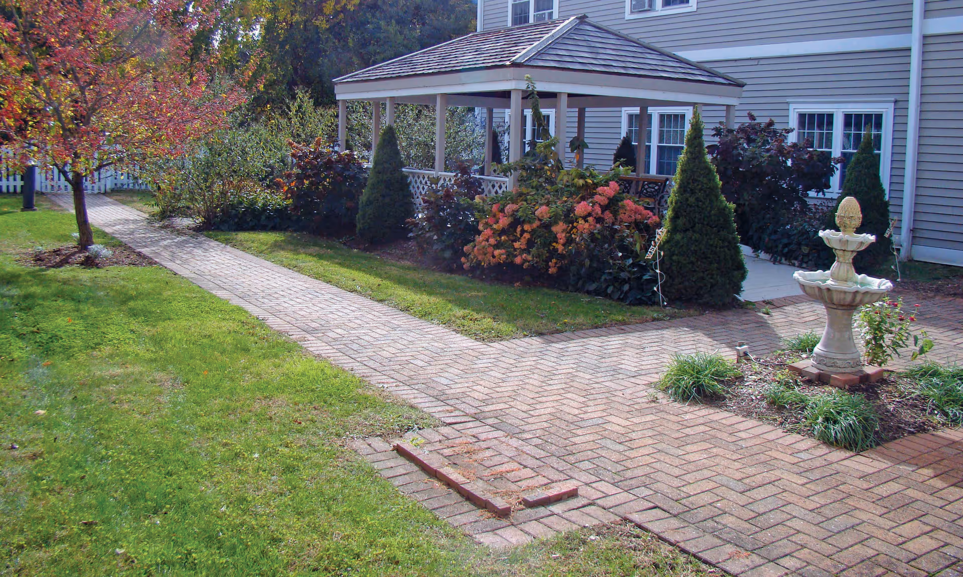A peaceful outdoor garden area at Apple Rehab Laurel Woods featuring a brick walkway, a white gazebo surrounded by bushes and flowers, a small fountain, and a tree with autumn-colored leaves. The building exterior with windows is visible in the background.