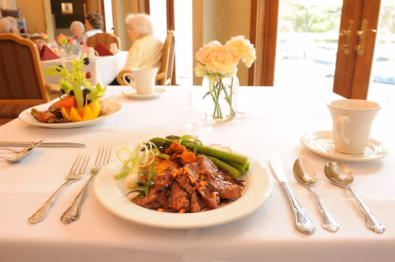 A dining table set with a white tablecloth, a plate of cooked meat with asparagus and garnishes, silverware, a cup and saucer, and a small vase with pink flowers. In the background, elderly people are seated at other tables near large windows with wooden frames.