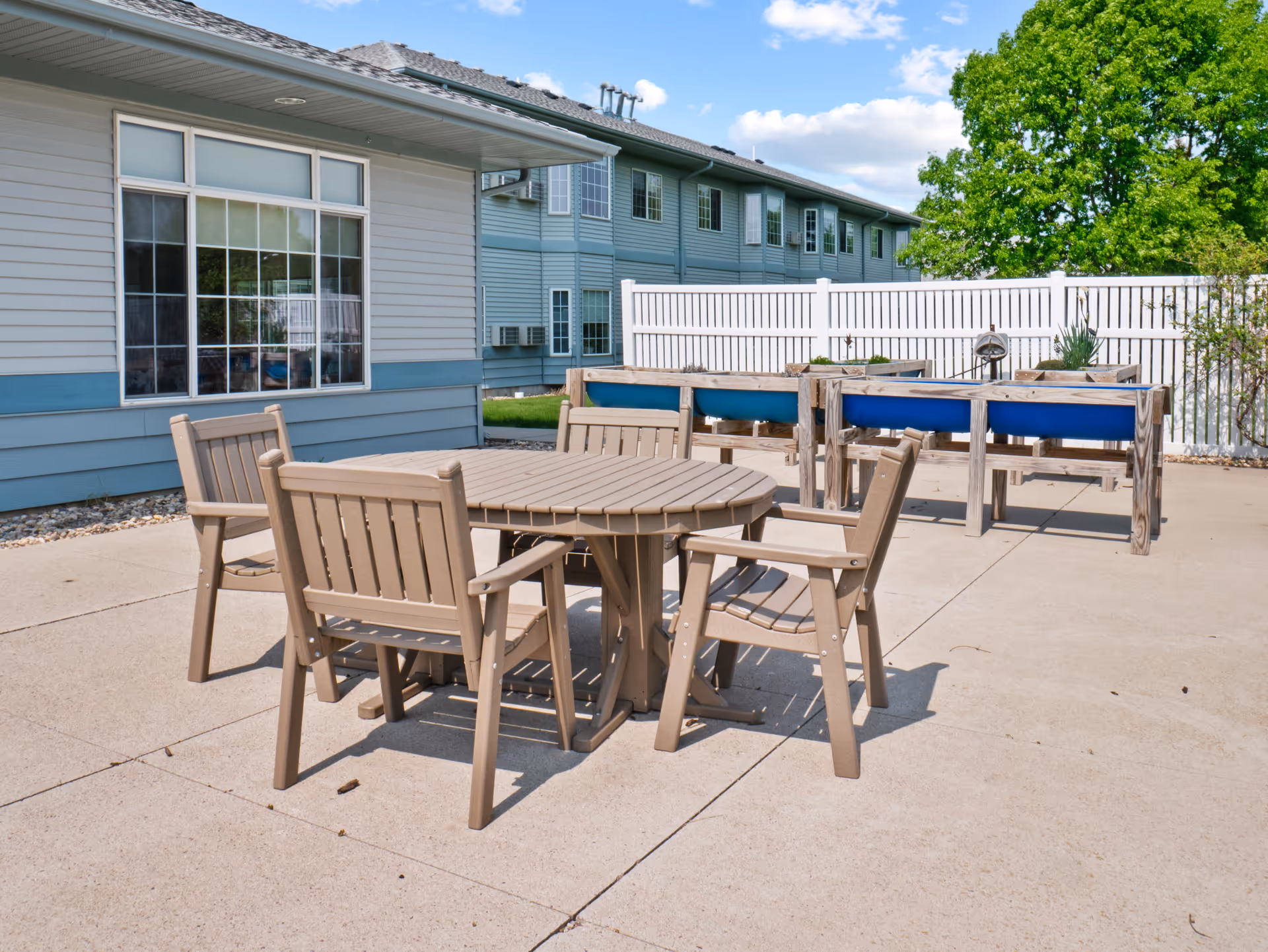 Outdoor patio area with a round table and four wooden chairs on a concrete surface. In the background, there are raised garden beds with plants and a white fence. The building exterior is visible with blue and gray siding and multiple windows. A green tree is also visible under a partly cloudy sky.
