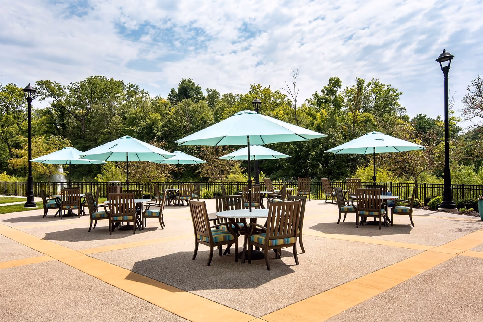 Outdoor patio area with multiple round tables and wooden chairs, each table shaded by a large light blue umbrella. The patio is surrounded by greenery and trees under a partly cloudy sky.