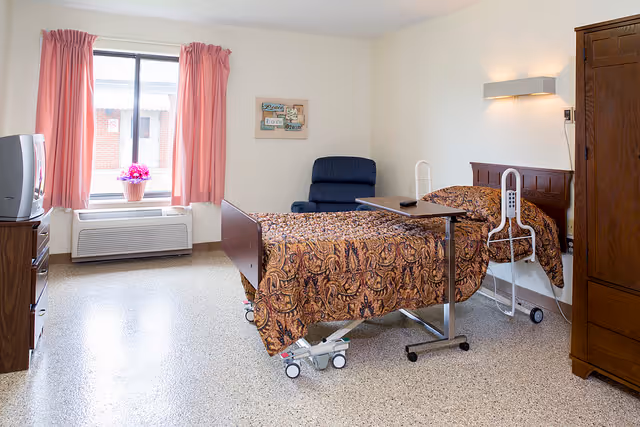 A senior living facility bedroom with a hospital-style bed covered in a patterned brown bedspread. There is a wooden headboard and a bedside table with a remote control. A blue recliner chair is positioned near the bed. The room has a window with pink curtains and a potted plant on the windowsill. A wooden wardrobe and a TV on a stand are also visible. The floor is speckled and the walls are light-colored.