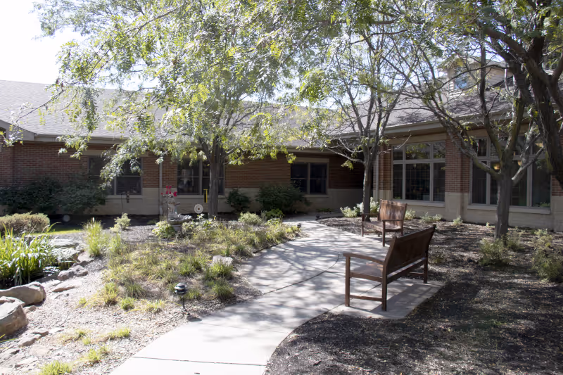 Outdoor courtyard area at Brookestone Village featuring a curved concrete walkway, two wooden benches, several trees providing shade, and landscaped garden beds with various plants and shrubs surrounding the path.