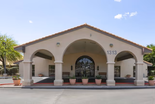 Front entrance of a single-story stucco building with an arched covered portico, potted plants, benches, and the address number 1313.