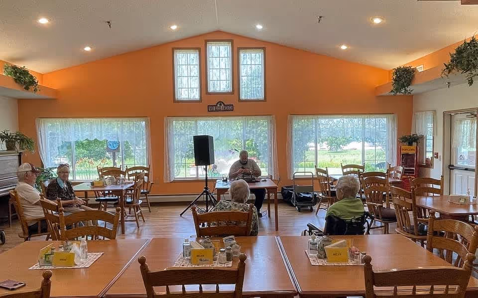 A bright dining room with orange walls and large windows letting in natural light. Several elderly people are seated at wooden tables and chairs, some facing a man sitting at a table with a microphone and speaker setup. The room has plants on shelves near the ceiling and a popcorn machine near the door.