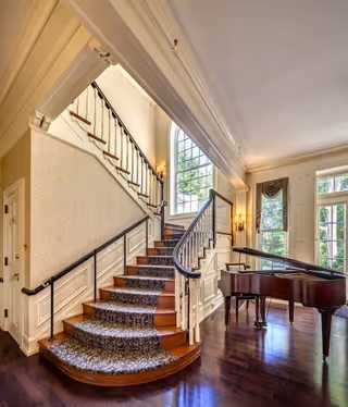 Interior view of a senior living facility featuring a curved wooden staircase with patterned carpet runner, white paneled walls, and black handrails. To the right of the staircase is a grand piano on polished dark hardwood floors. Large windows with decorative curtains allow natural light to fill the room.