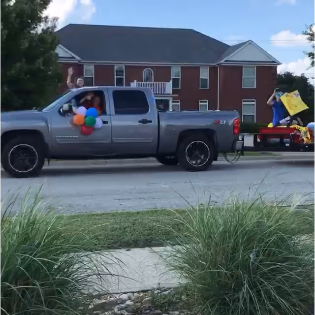 A silver pickup truck decorated with colorful balloons on the driver's side door is driving on a street in front of a two-story brick building with white trim. A person inside the truck is waving, and another person in the truck bed is holding a yellow bag. Green bushes and grass are visible in the foreground under a partly cloudy sky.