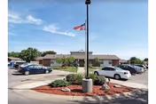 Circular driveway and parking lot with a flagpole in front of a single-story Prairie Villa building under a blue sky.