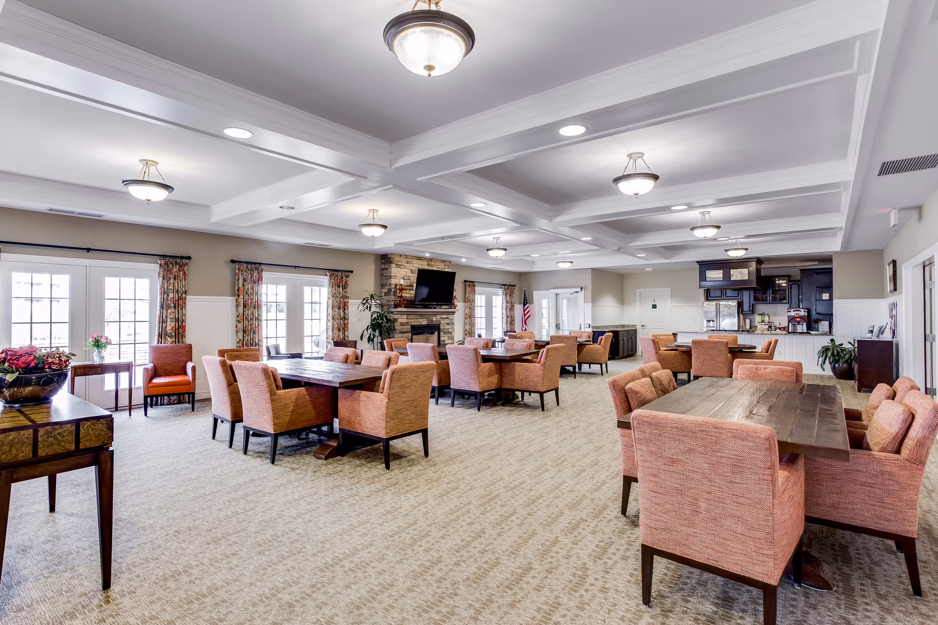 A spacious, well-lit common area with multiple wooden tables surrounded by orange upholstered chairs. The room features a coffered ceiling with recessed lighting and hanging light fixtures. There are large windows with floral curtains letting in natural light, a stone fireplace with a mounted flat-screen TV, and a kitchenette area with dark cabinetry and stainless steel appliances in the background.