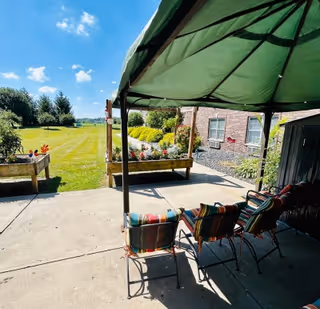 Shaded patio with colorful cushioned chairs under a green canopy overlooking a grassy lawn and gardens beside a brick building.