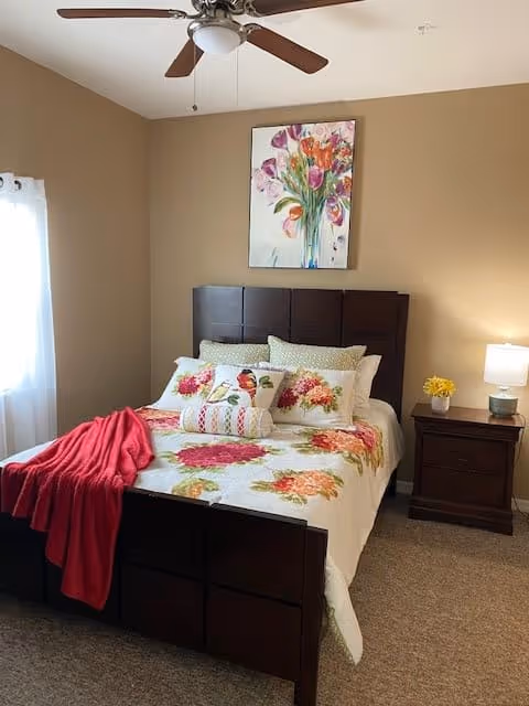 A cozy bedroom with a dark wooden bed frame and a floral bedspread featuring red and pink flowers. The bed is adorned with multiple pillows and a red throw blanket. A wooden nightstand with a small lamp and a yellow flower arrangement is beside the bed. A ceiling fan with wooden blades is mounted on the ceiling, and a window with white curtains allows natural light into the room. A colorful floral painting hangs on the beige wall above the bed.