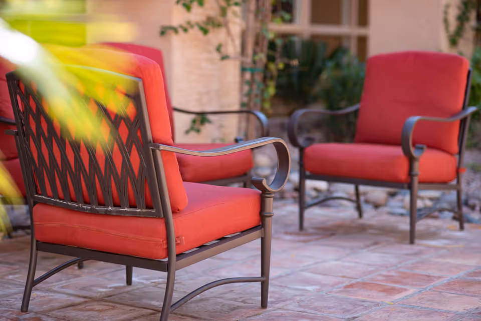 Two outdoor chairs with red cushions on a tiled patio with some greenery and a building wall in the background.