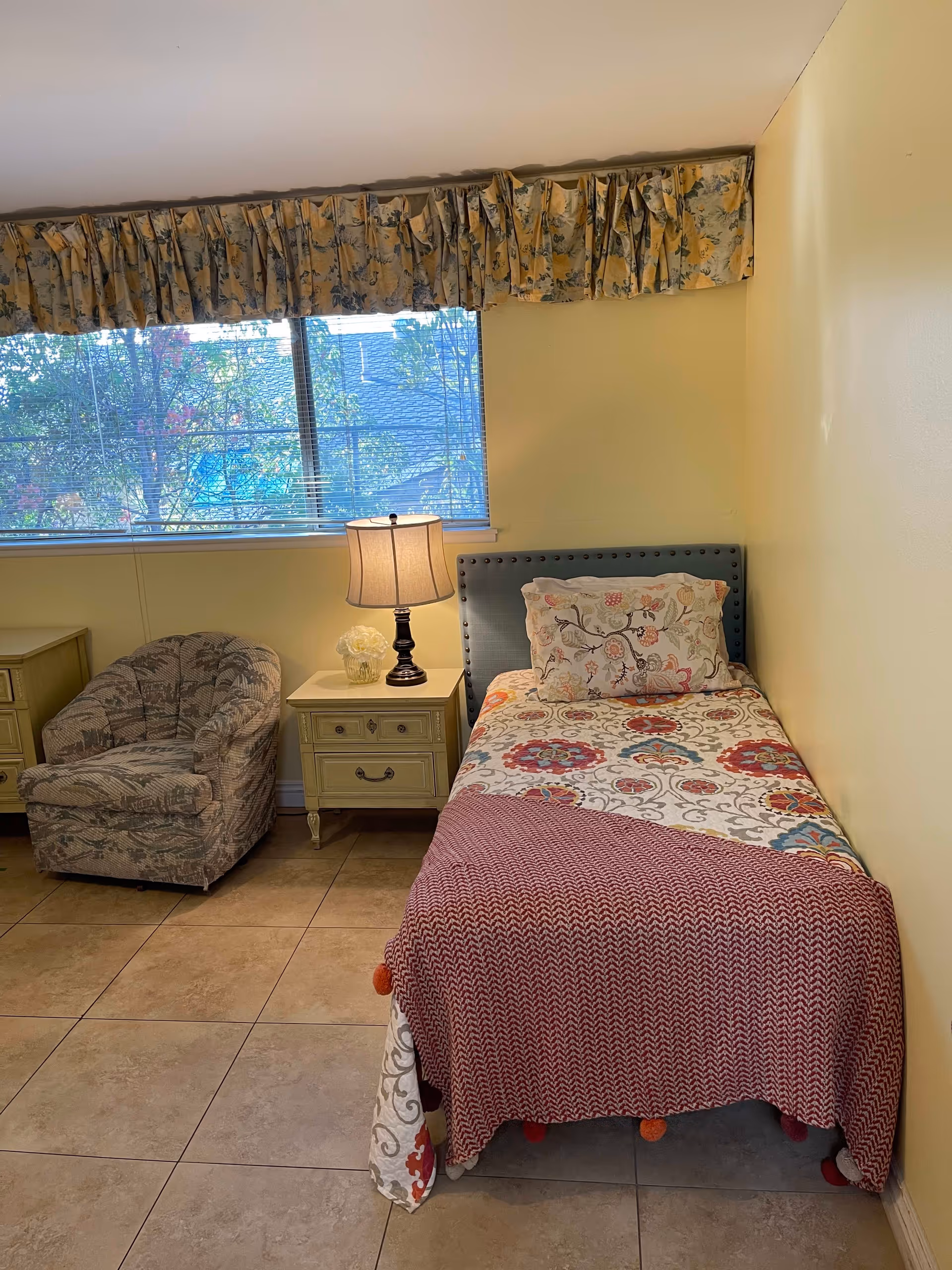 A small bedroom with a single bed covered in a colorful floral bedspread and a red knitted blanket. Next to the bed is a light yellow nightstand with a lamp and a small flower arrangement. There is a cushioned armchair with a patterned fabric to the left of the nightstand. A large window with floral valance curtains lets in natural light, and the floor is tiled.