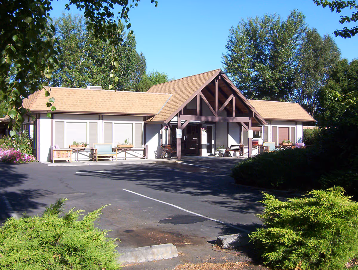Single-story building with a covered entrance, outdoor benches, and landscaping surrounded by trees under a clear blue sky.