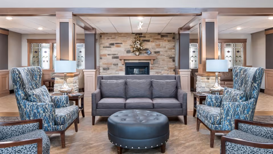 A cozy living room area with a gray three-seater sofa centered in front of a stone fireplace. Two blue patterned armchairs with matching pillows face the sofa, with a round black tufted ottoman in the middle. The room features wooden columns, side tables with lamps, and stained glass windows in the background.