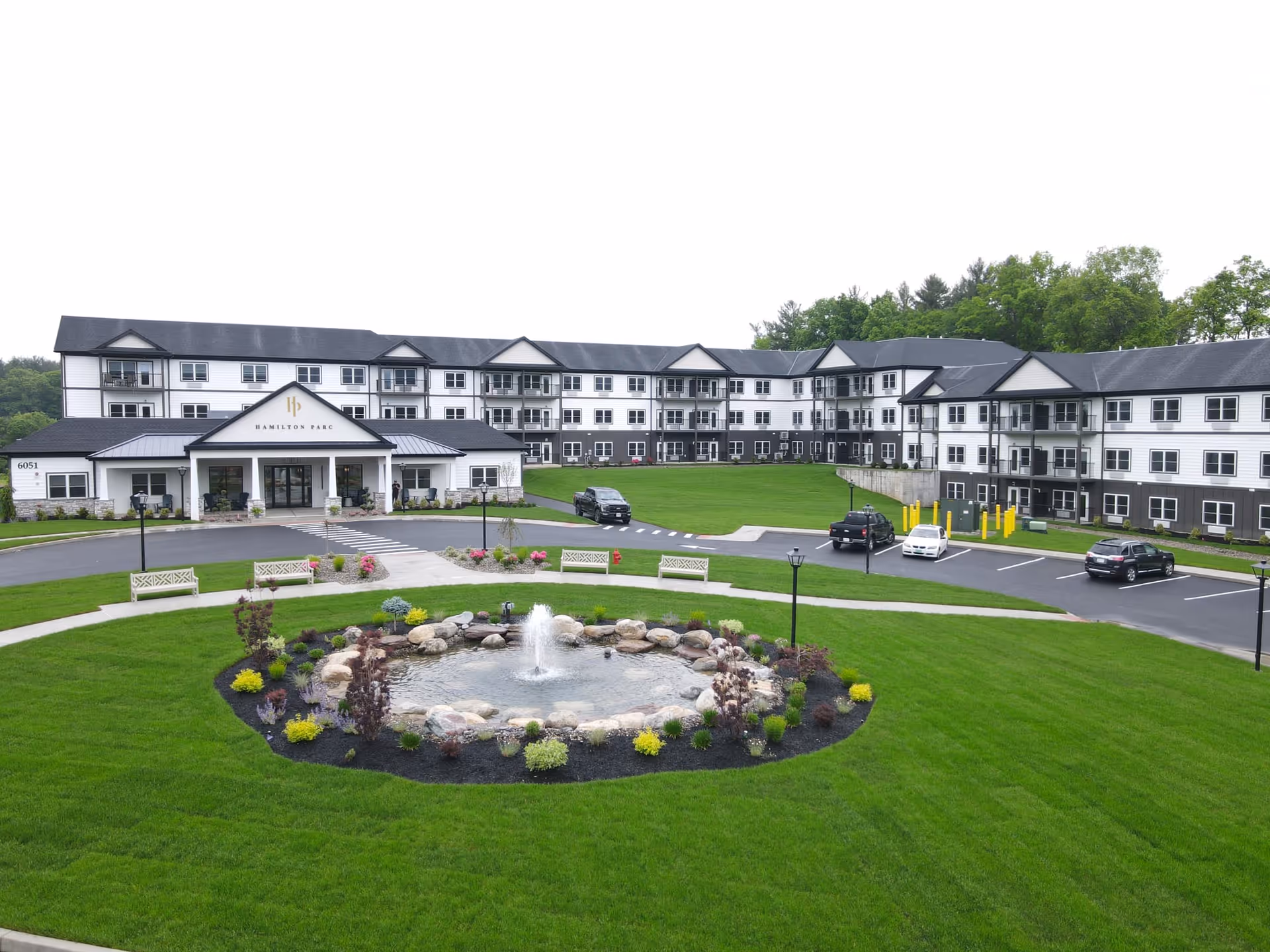 Exterior view of Hamilton Parc senior living facility showing a large three-story building with white and dark gray siding, multiple balconies, and a covered entrance. In front of the building is a circular landscaped area with a small pond and fountain surrounded by rocks and plants. There are benches and a paved driveway with parked cars. Trees are visible in the background.