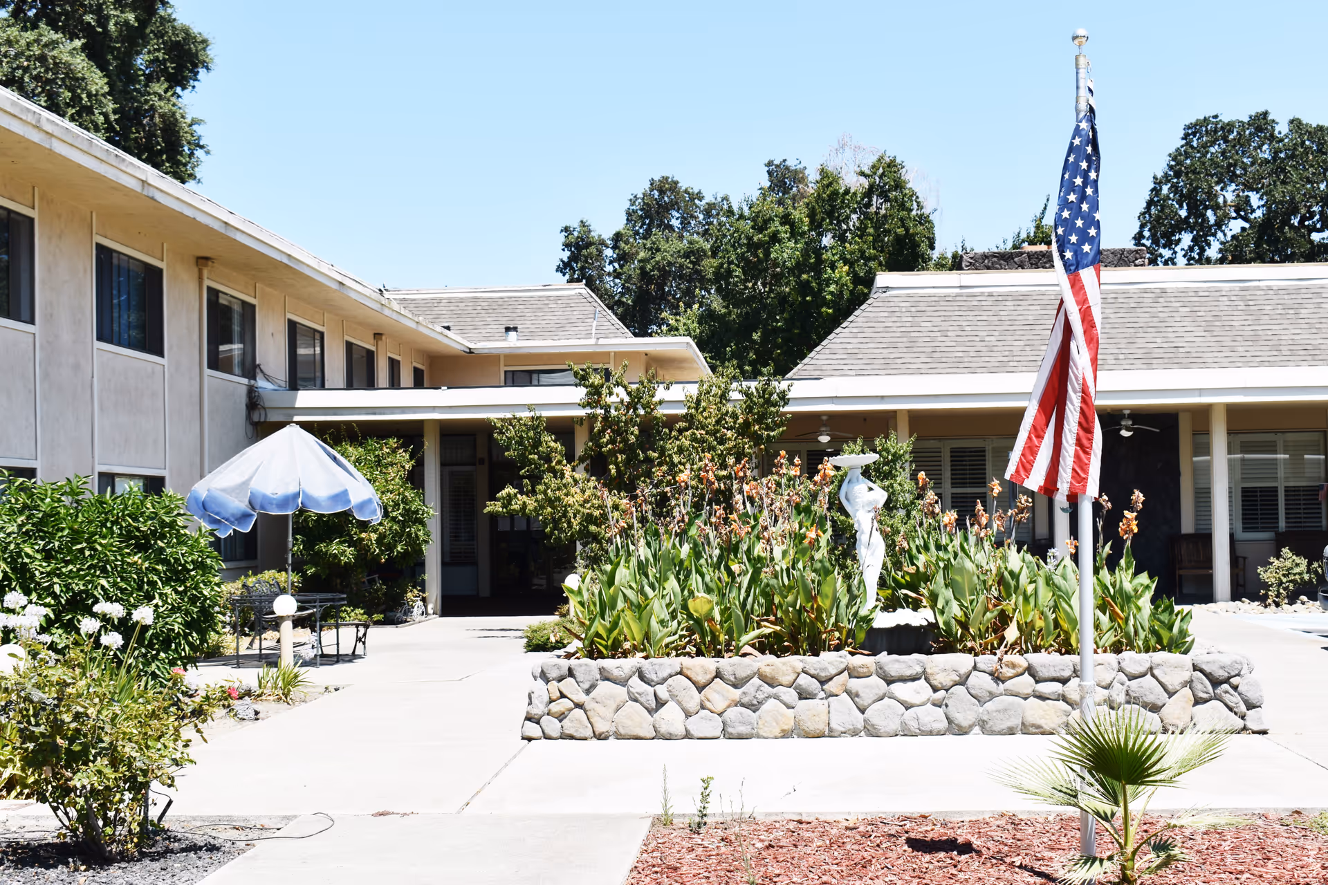 Outdoor courtyard area of Del Monte Assisted Living & Memory Care - Stockton featuring a stone planter with green plants and a white statue, an American flag on a flagpole, surrounding buildings with windows, and a blue umbrella shading a seating area.