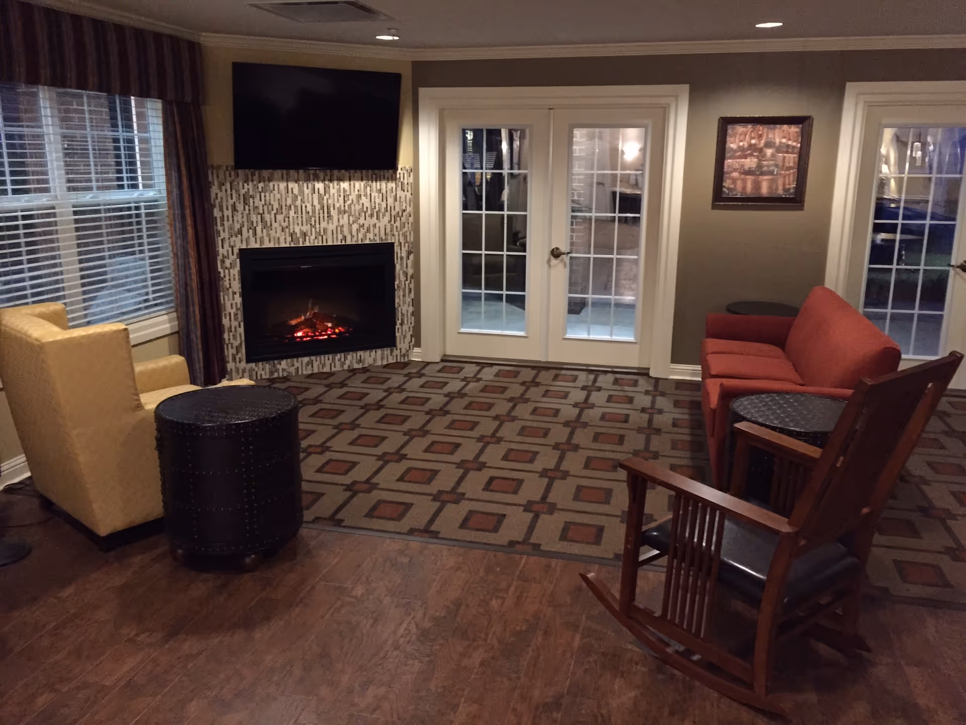 Cozy common room with a fireplace and wall-mounted TV, seating including armchairs, a sofa and a rocking chair, and glass French doors.