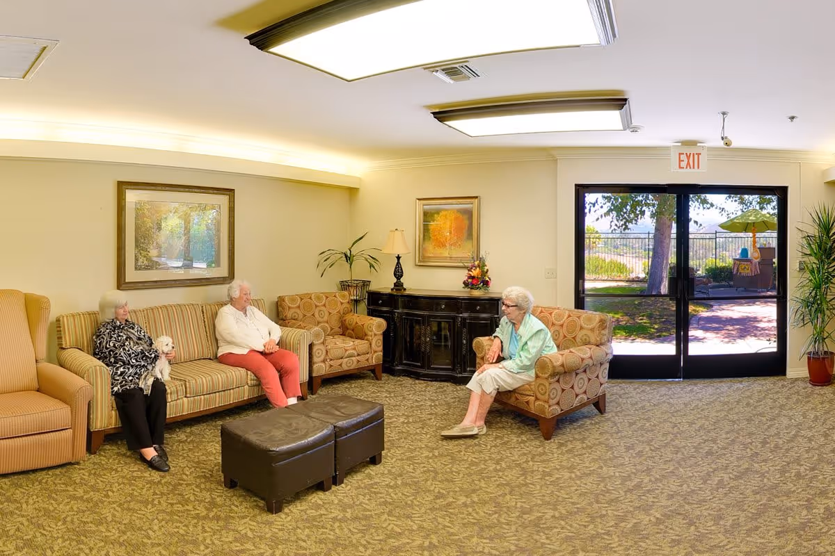 A senior living facility common area with three elderly women sitting on patterned armchairs and a striped sofa. One woman is holding a small white dog. The room has carpeted flooring, framed artwork on the walls, a black cabinet with a lamp and flower arrangement, and large glass doors leading to an outdoor garden area with trees and an umbrella-covered table.