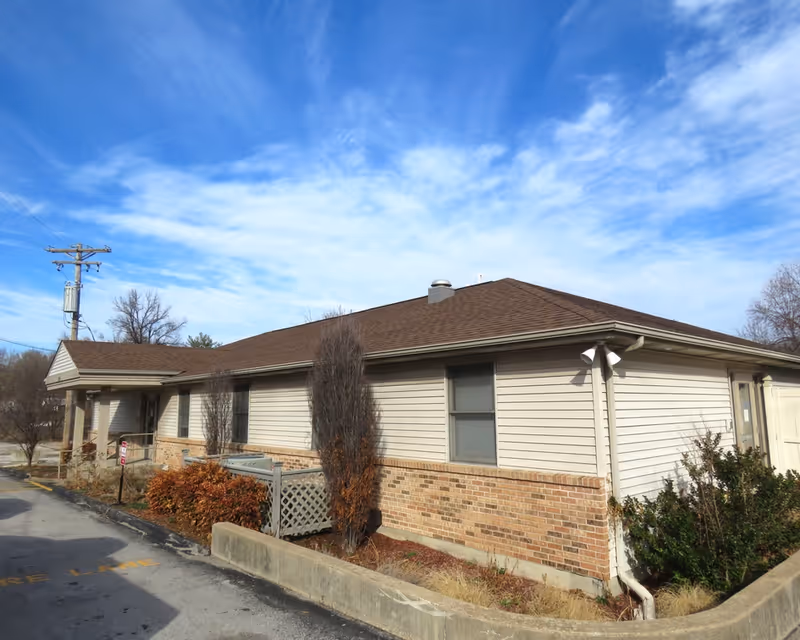 Exterior view of a single-story building with beige siding and brick accents under a partly cloudy blue sky. There are some leafless shrubs and bushes around the building, and a paved driveway or parking area is visible in the foreground.