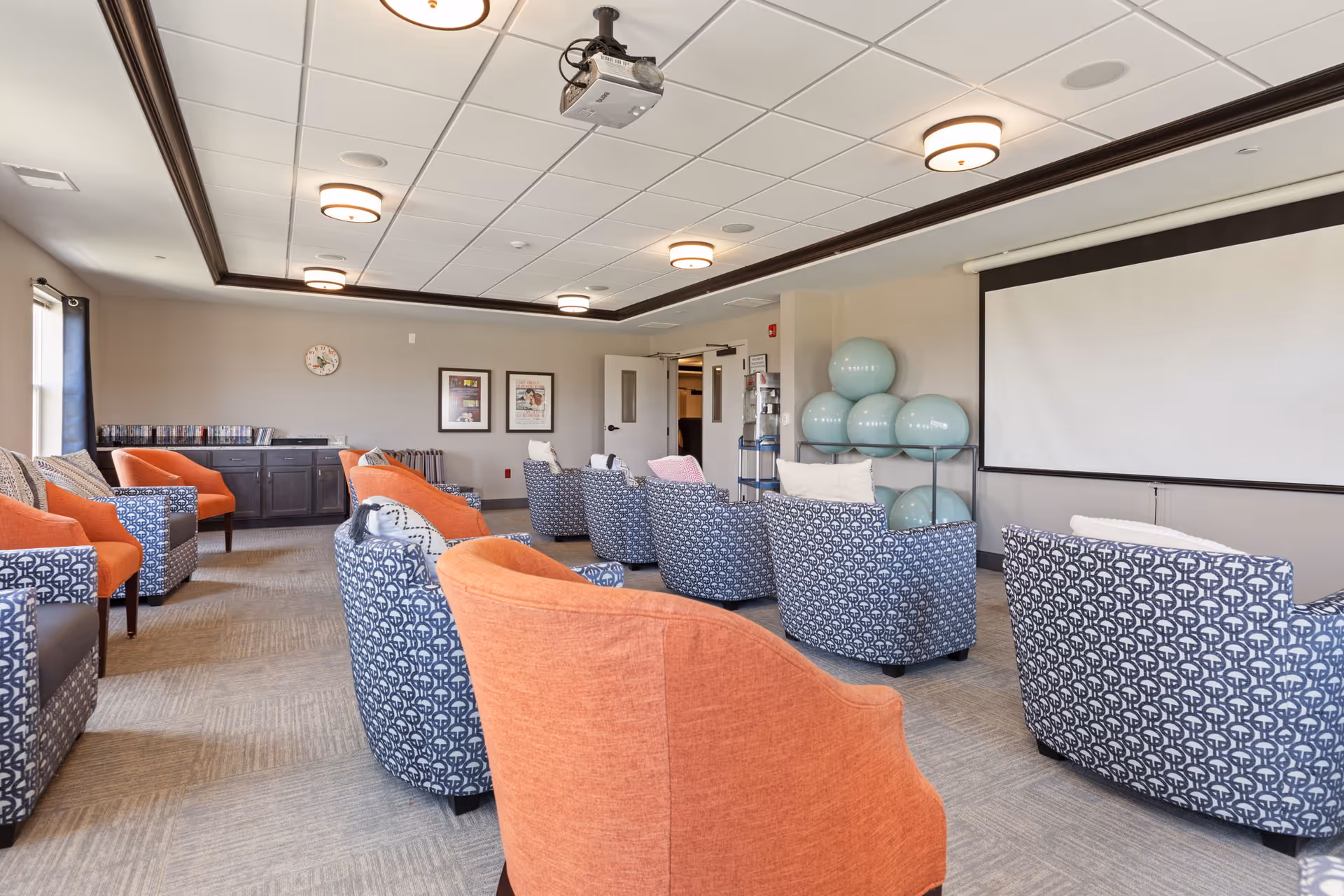 Bright common room with patterned and orange armchairs arranged facing a projection screen and stacked exercise balls.